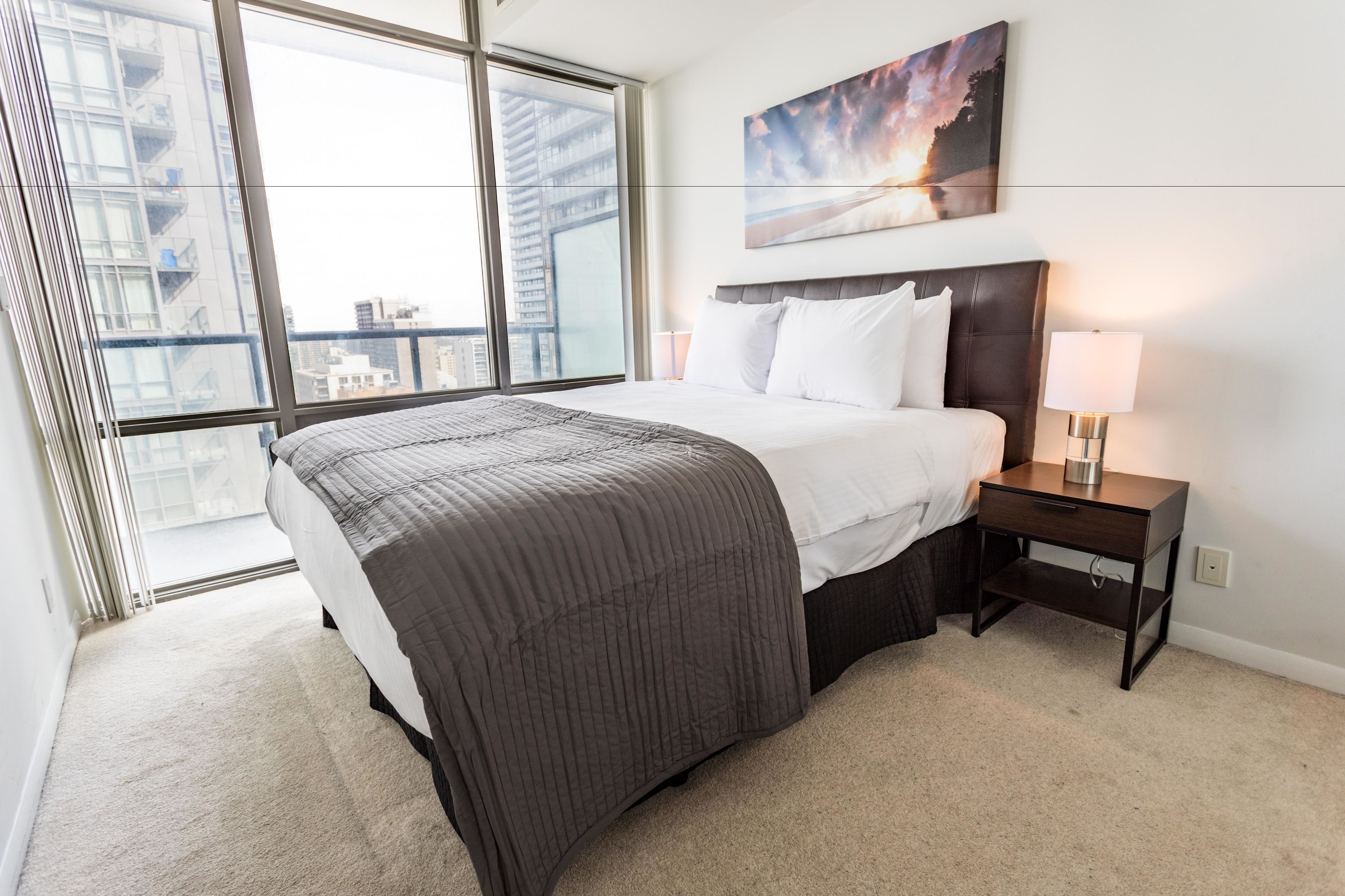Een moderne hotelkamer met een groot bed, een nachtkastje met lamp en uitzicht op de stad door raamhoogte ramen.