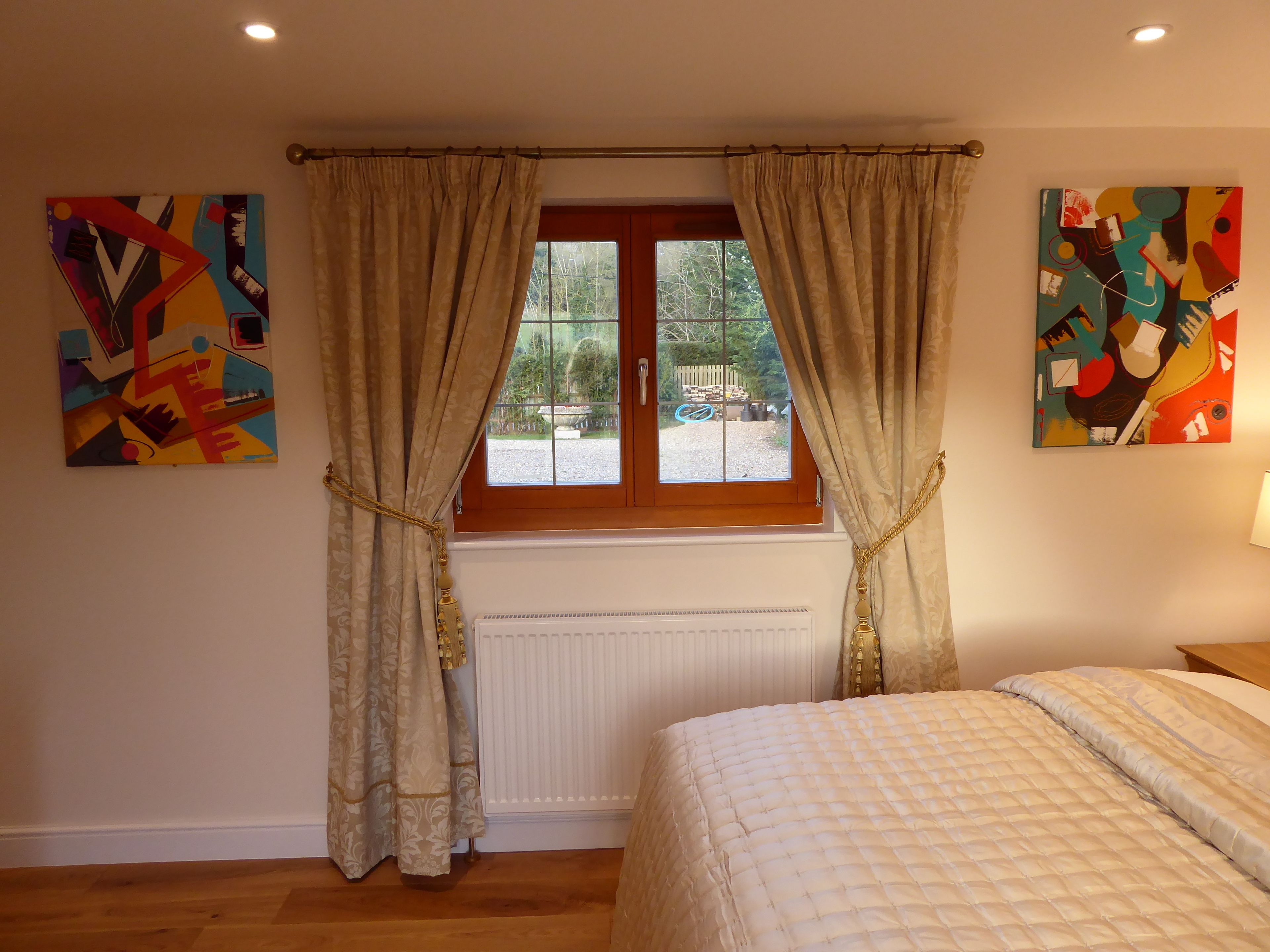 Un dormitorio con cama, ventana con cortinas y dos cuadros abstractos en las paredes.