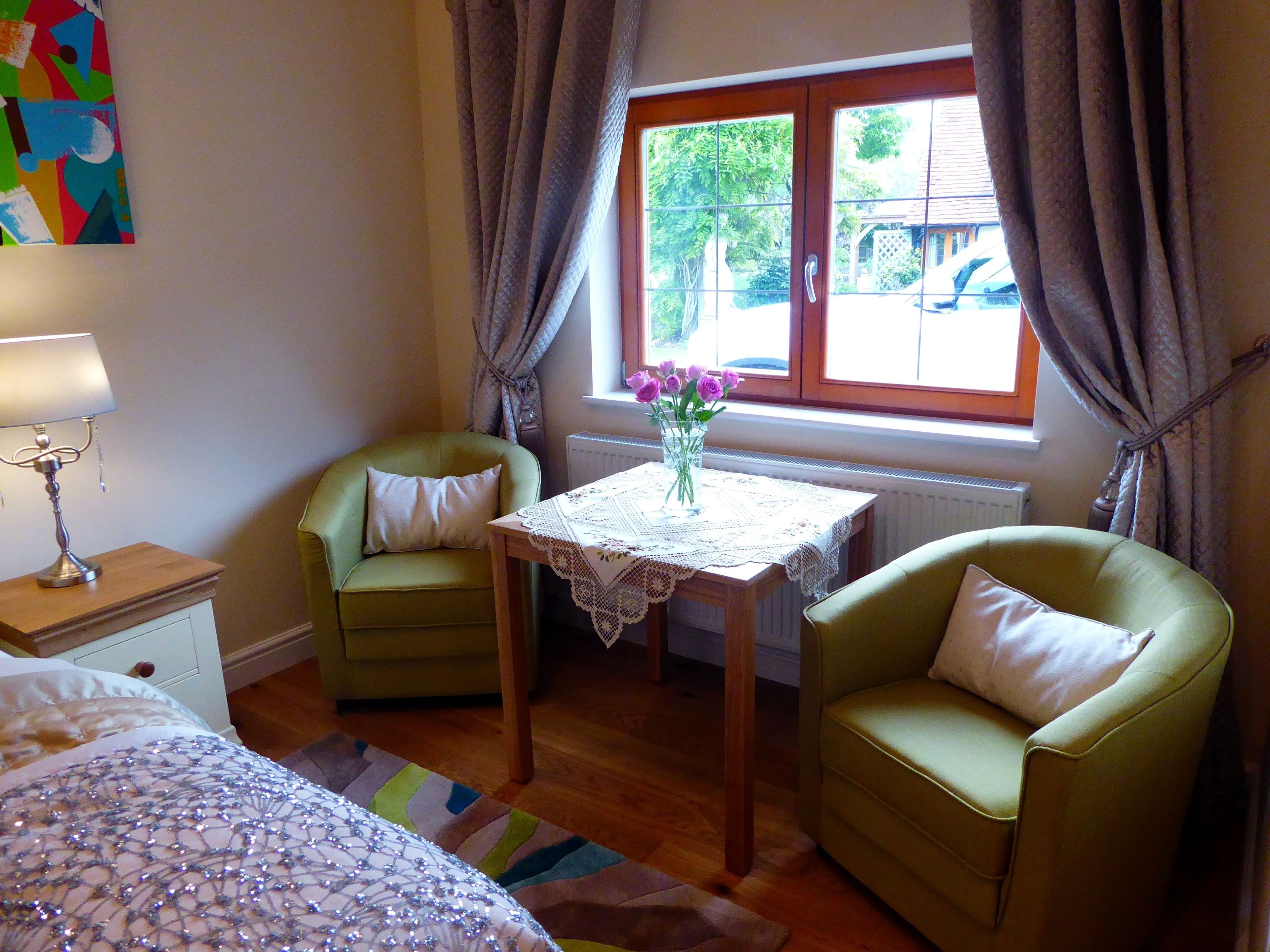 Una habitación pequeña y acogedora con dos sillones verdes, una mesita con mantel de encaje, una lámpara y una ventana con cortinas.