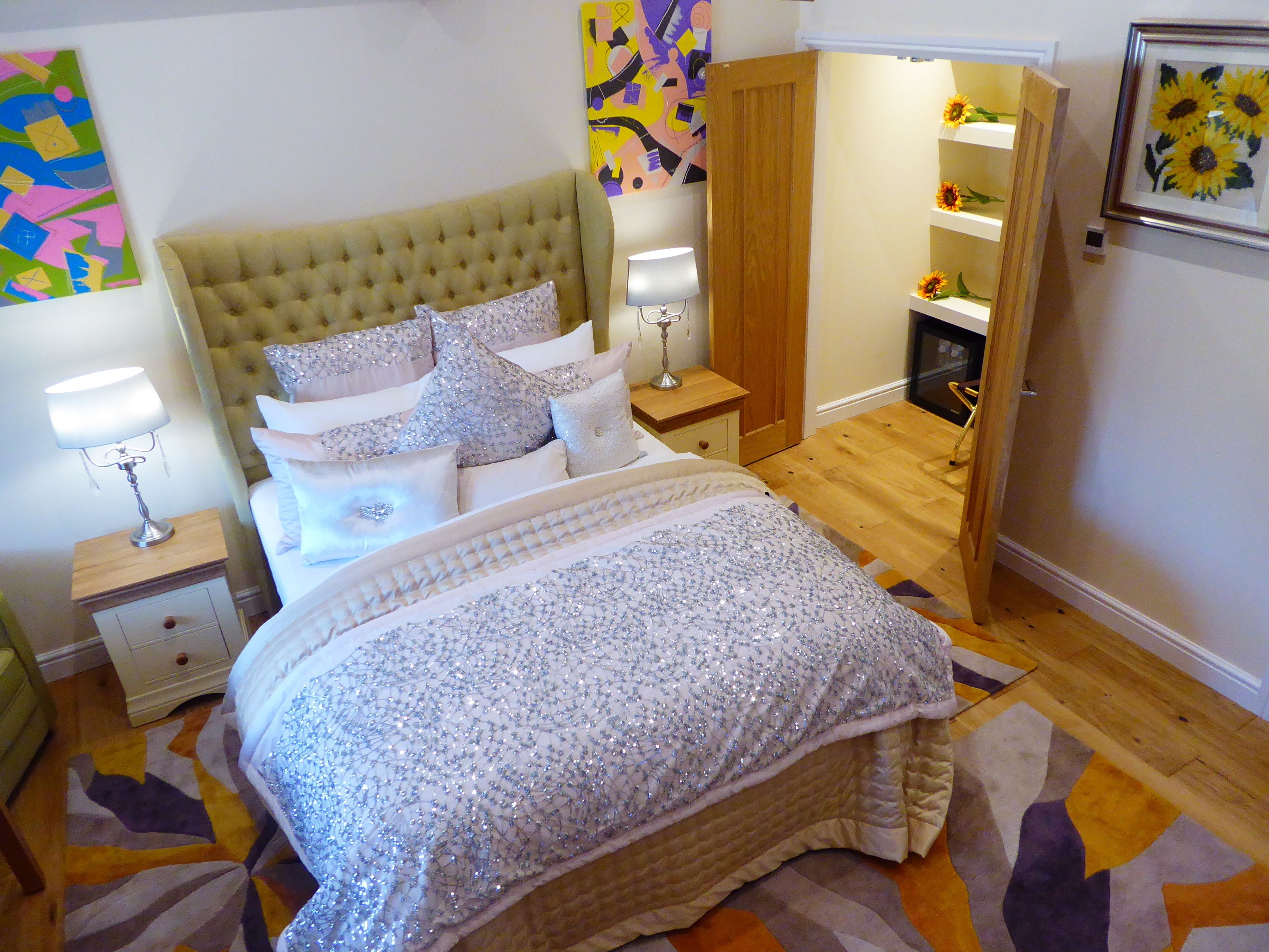 Une chambre à coucher avec une tête de lit capitonnée, un lit recouvert d’un tissu à paillettes, une table de chevet avec une lampe et un plancher en bois.