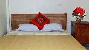 Una cama con cabecera de madera, dos almohadas blancas y una almohada decorativa roja. A la derecha, una mesita de noche con un jarrón de flores rojas.