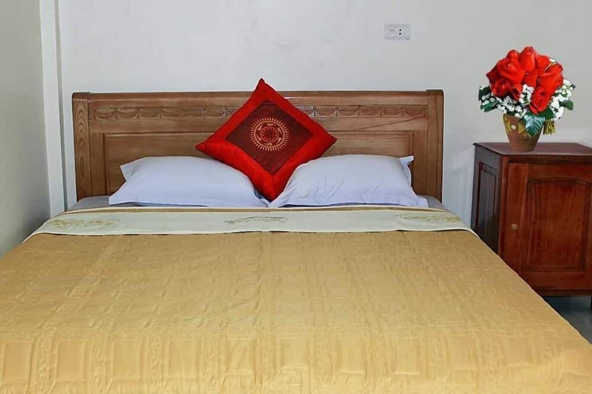 Ein Bett mit Holz-Kopfstück, zwei weiße Kissen und ein rotes Zierkissen. Dazu ein Nachttisch aus Holz mit einer Vase mit roten Blumen.