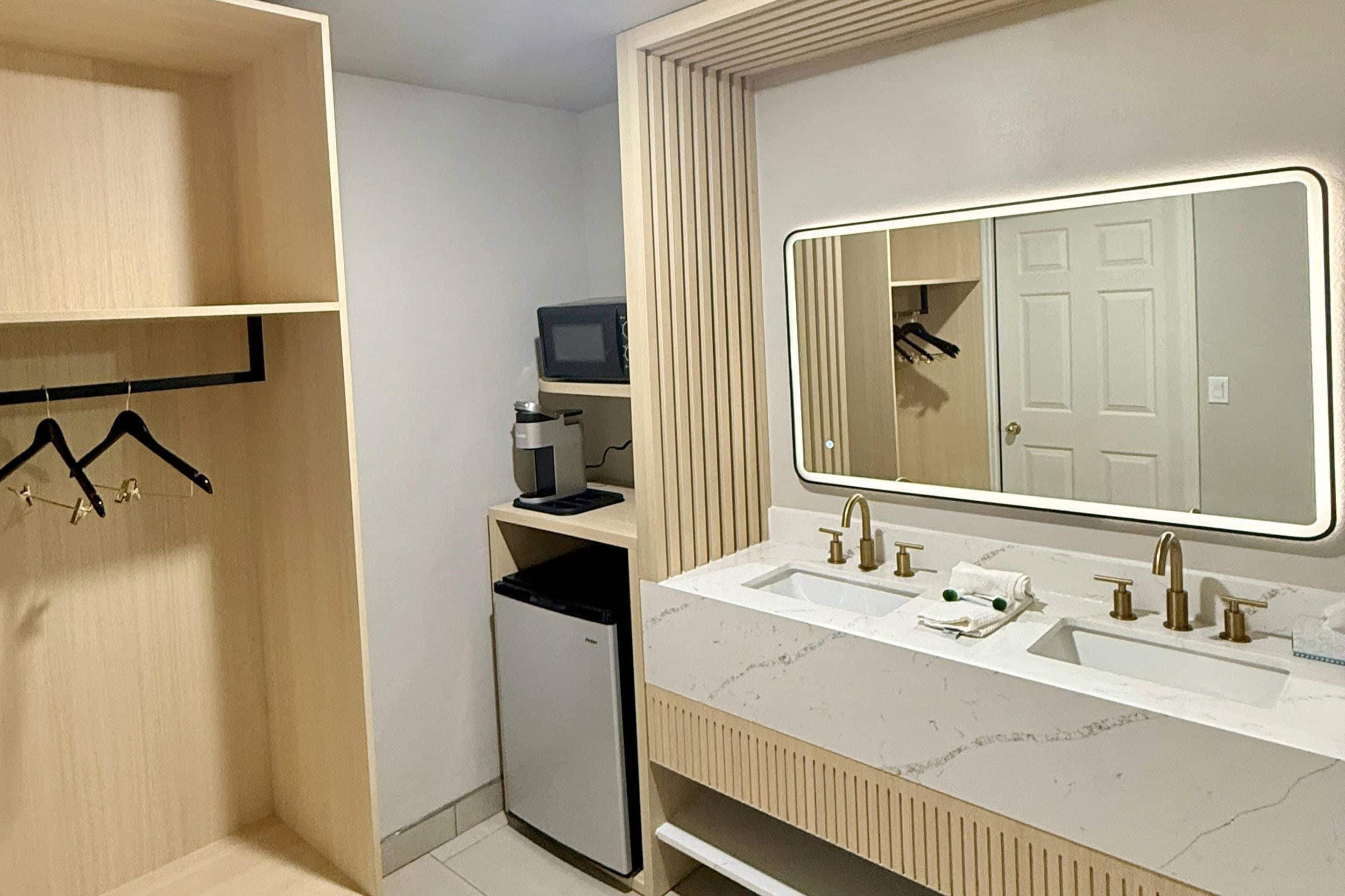 A modern bathroom with a double sink, a mirror with a light, and a small refrigerator.