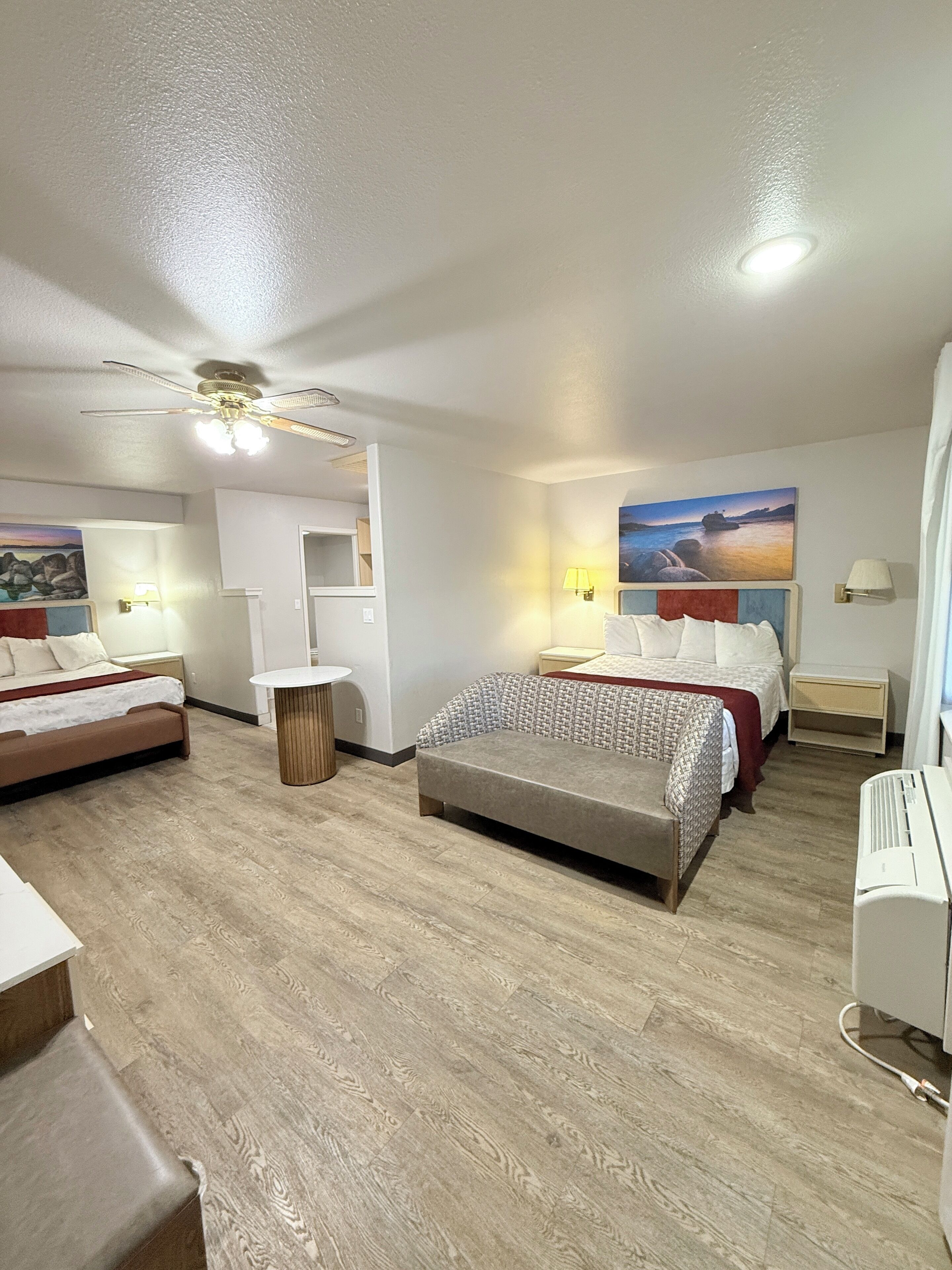 Habitación de hotel con dos camas, ventilador de techo, un cuadro de un paisaje y una mesita.