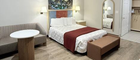 Habitación de hotel con cama, sofá, una pequeña mesa, una banca, un cuadro, un espejo y una puerta.