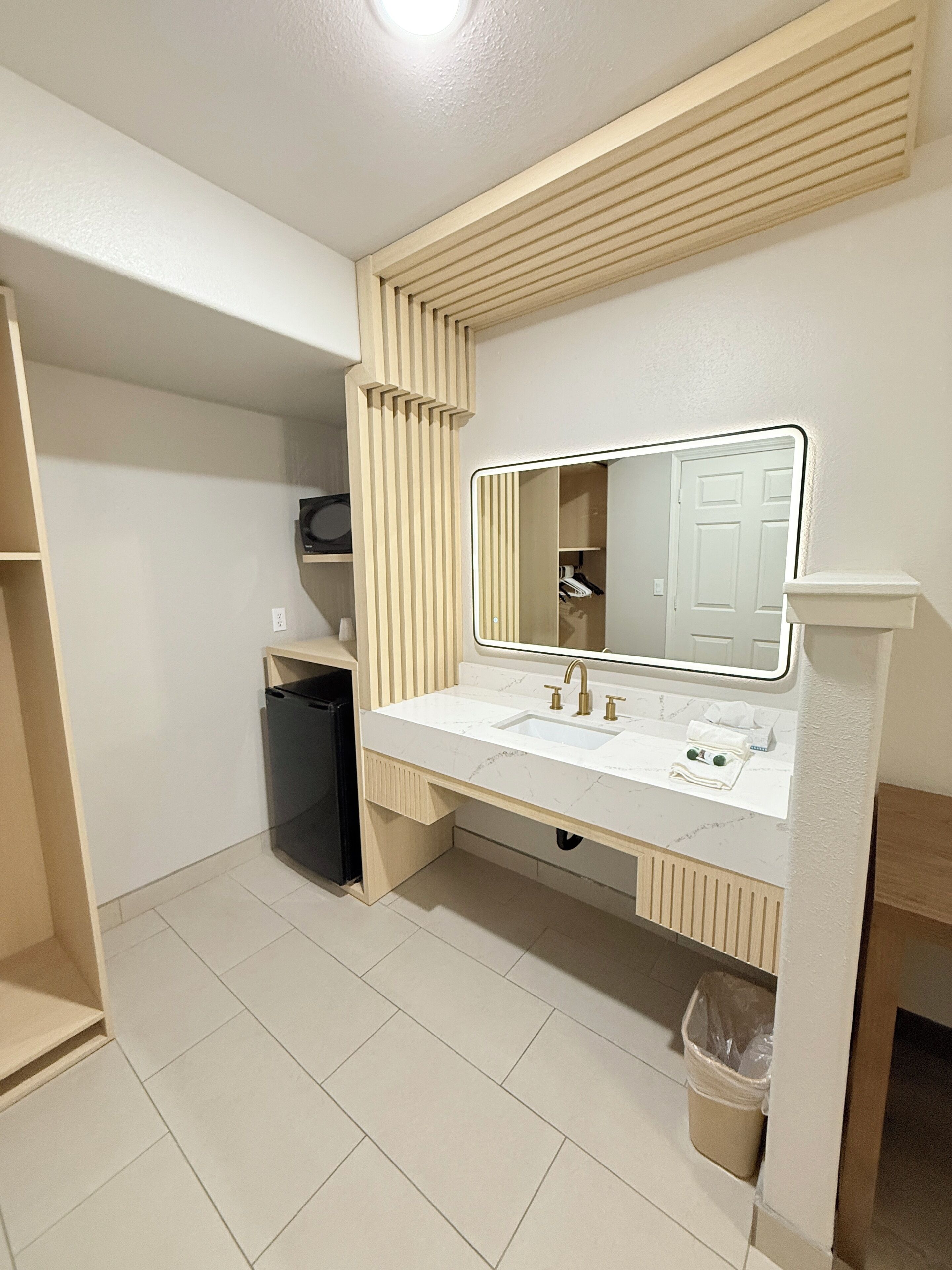 A modern bathroom with a white sink, mirror, and vanity. There is a trash can and a towel rack visible.