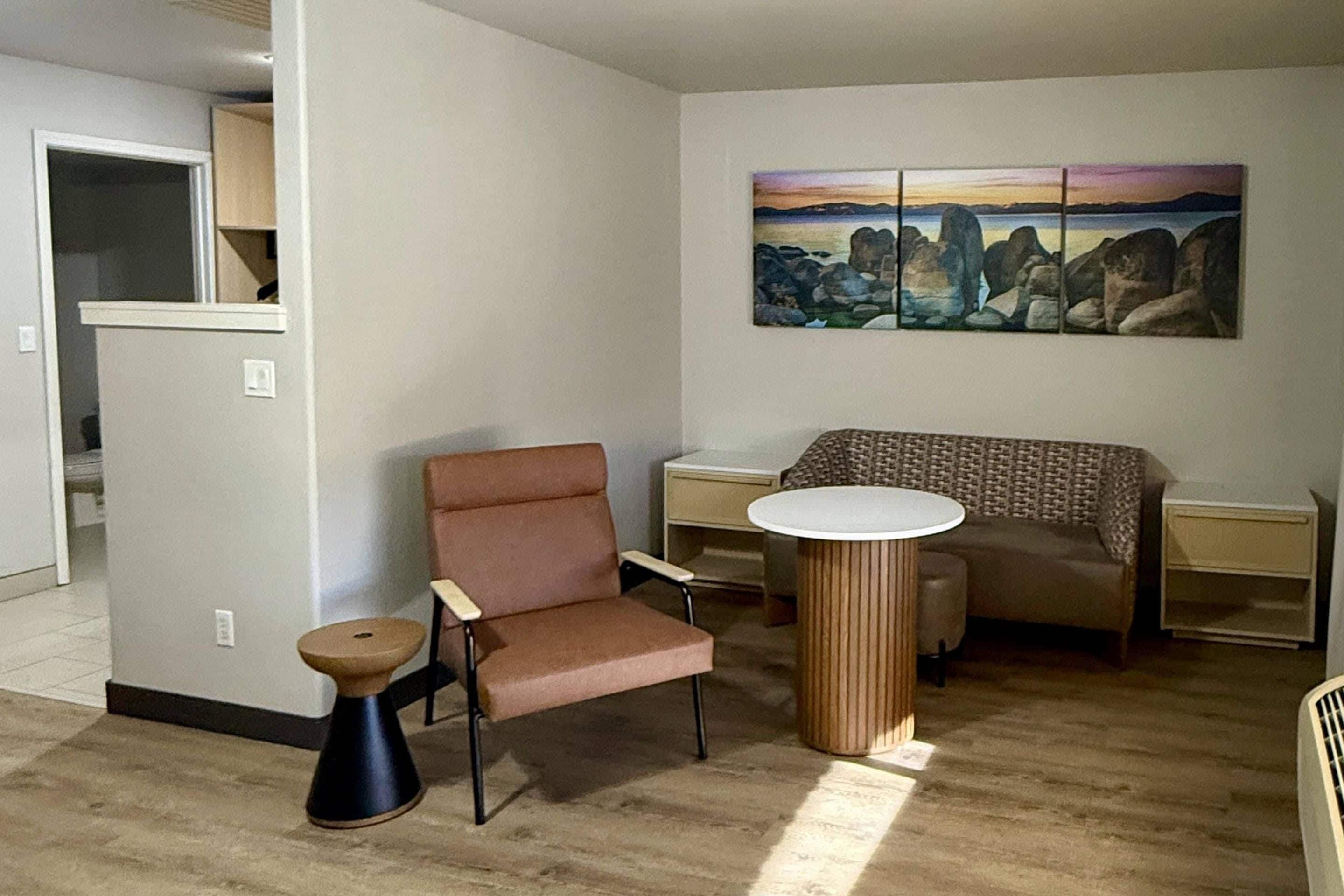 A living room with a brown armchair, a round table, a small stool, and a painting on the wall.