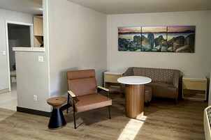 Una sala de estar con un sillón marrón, una mesa redonda, un taburete pequeño y un cuadro en la pared.