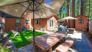 A backyard with a wooden table and benches, surrounded by a patio area with a large umbrella. - Breezy Pines Cabin | Backyard Picnic | BBQ (South Lake Tahoe)