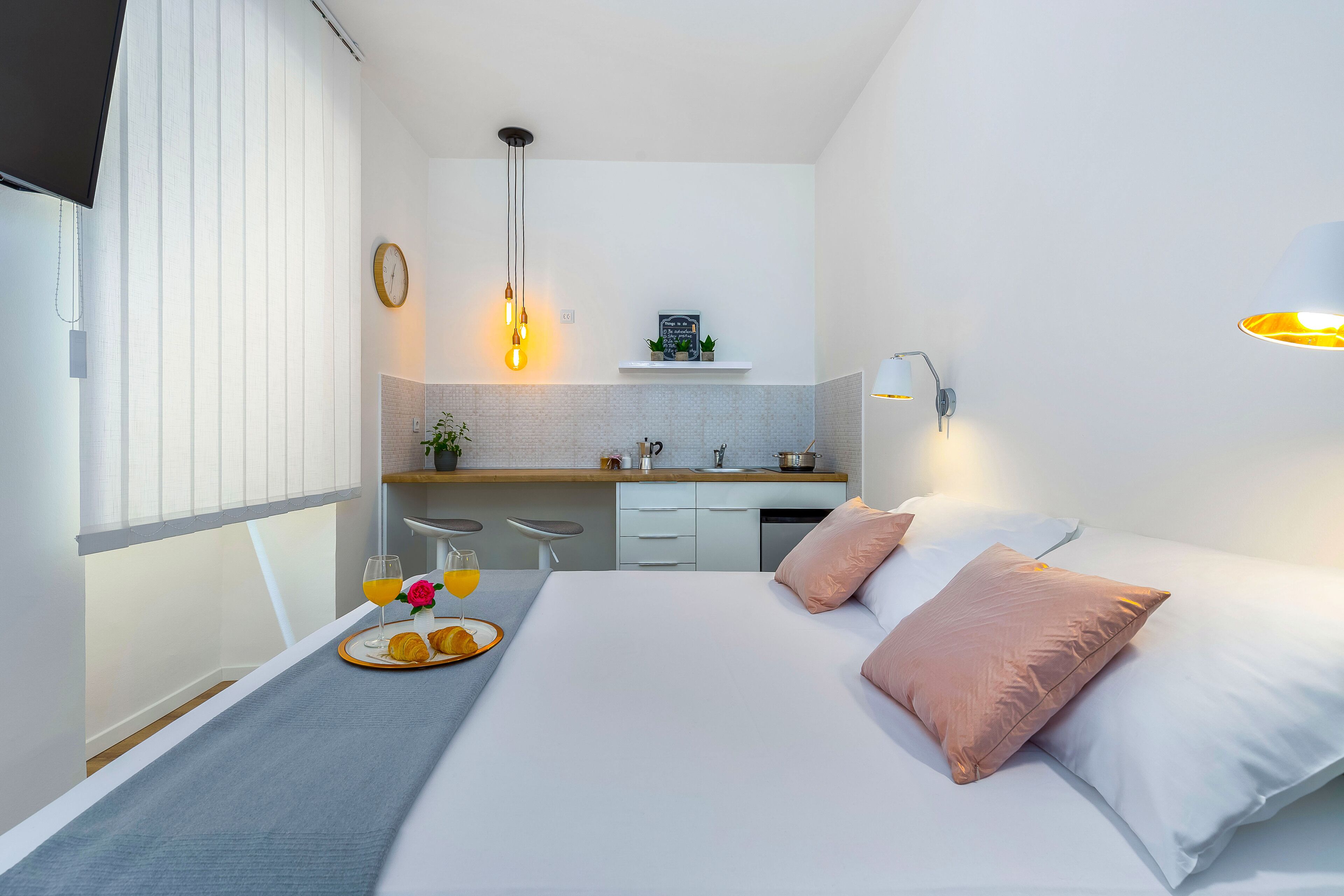 Une chambre d’hôtel moderne équipée d’un lit, d’une table de chevet avec des articles pour le petit-déjeuner, d’un petit coin cuisine et d’une fenêtre avec des stores.