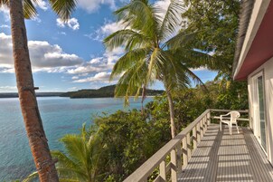 A wooden deck with a white chair, overlooking a turquoise sea and palm trees.