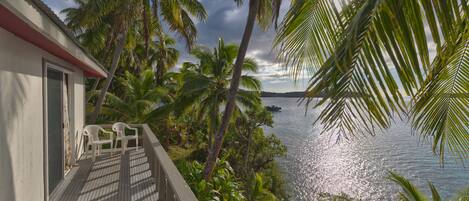A wooden deck with two white chairs, overlooking a body of water with palm trees and a building with a red roof.