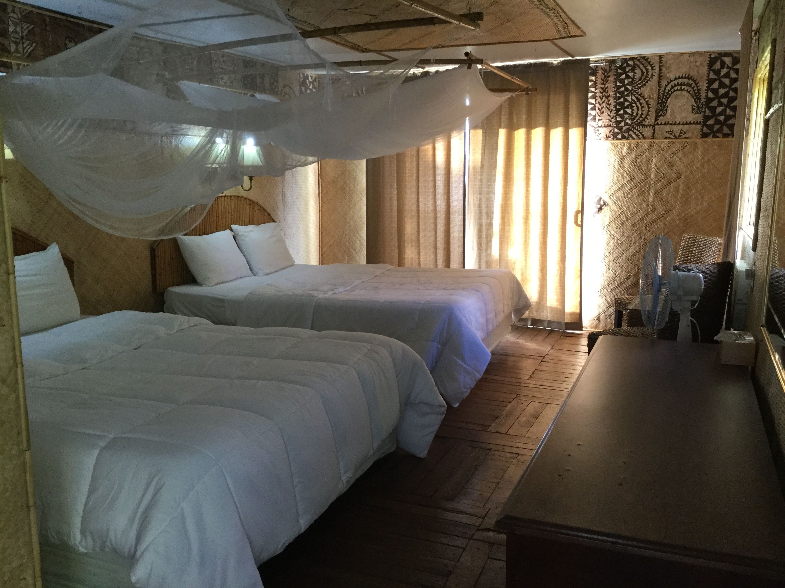 A room with two beds, a wooden floor, a small table, and a fan.