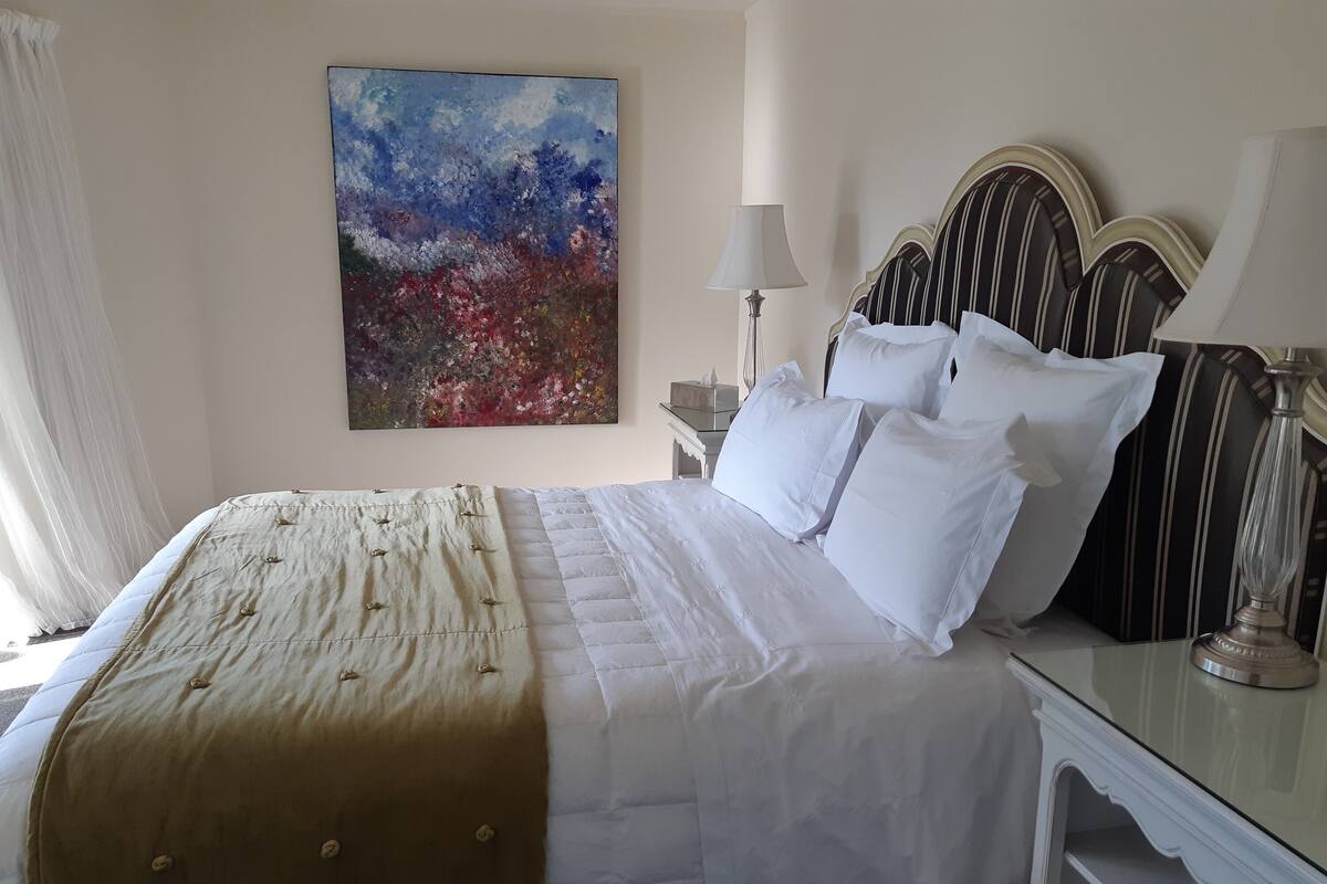A neatly made bed with white linens and a beige throw blanket, a bedside table with a lamp, and a painting on the wall.