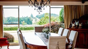 A dining room with a large wooden table, six chairs, a chandelier, and a view of a garden through large windows.