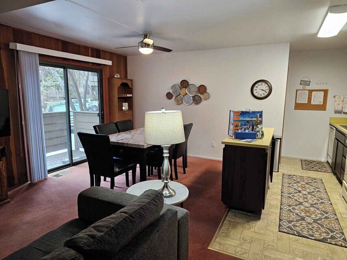 A living room with a sofa, a dining table with chairs, a lamp, a clock, and a small kitchen area with a sink and counter.