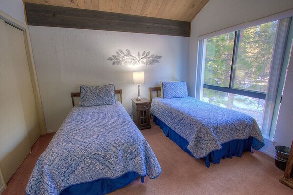 Une chambre avec deux lits, une table de chevet, une lampe et une fenêtre donnant sur des arbres.