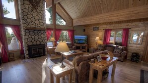 Interior acogedor de una cabaña de troncos con chimenea de piedra, muebles de madera y ventanas amplias.