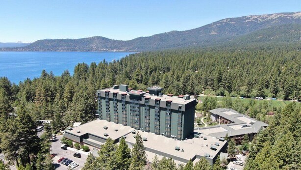 A modern hotel with a central building surrounded by a forested area, a parking lot, and a view of a lake and mountains.