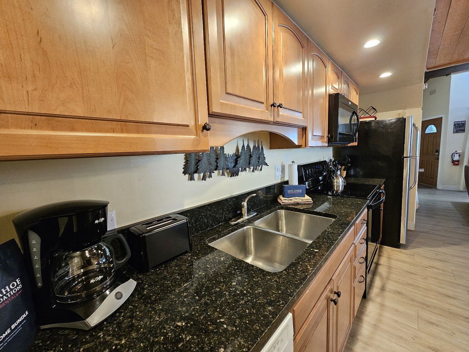 A kitchen with wooden cabinets, a stainless steel sink, a black countertop, and a black refrigerator.