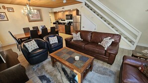 A spacious living room with a brown leather sofa, black leather armchairs, a wooden coffee table, and a dining area with a wooden table and chairs. A staircase is visible in the background leading to an upper level.