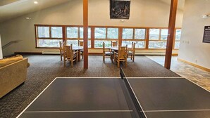 A room with a ping pong table, wooden chairs, a couch, and large windows.