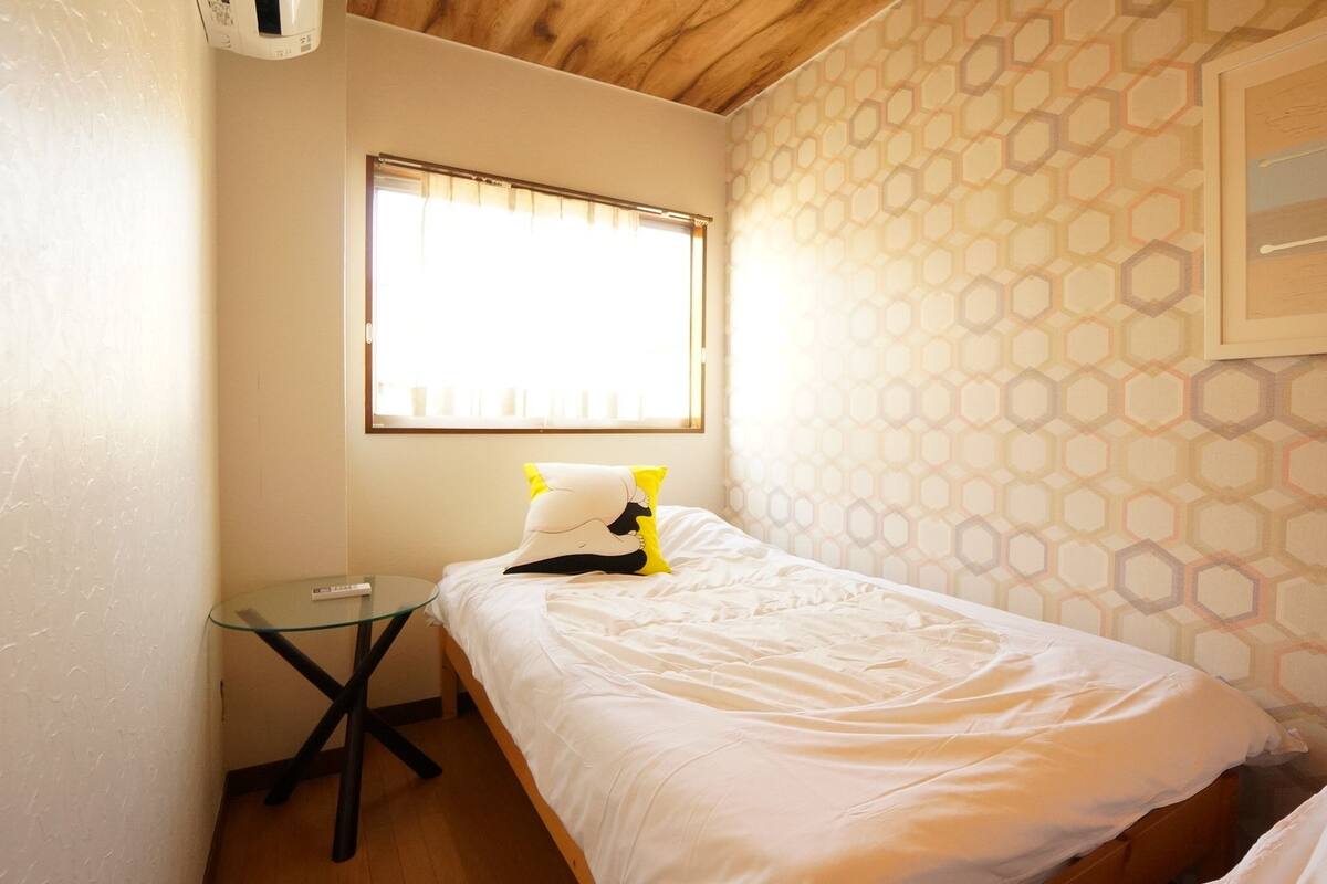 A small, neatly made bed with white bedding and a yellow pillow, a glass table with a remote control, and a window with blinds.