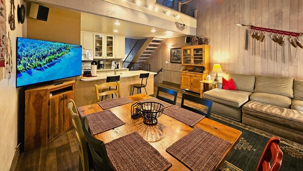 A spacious living area with a wooden dining table, a sofa, and a TV. A kitchen is visible in the background.
