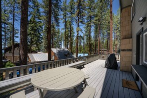 A wooden deck with a table and benches, surrounded by tall trees and a building with a sloped roof. - Walk to Beach | Pet-Friendly | Deck | BBQ (Zephyr Cove)