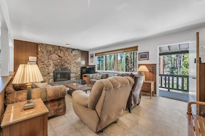 Una sala de estar con pared de piedra, un sofá, un sillón reclinable, una mesa de centro, una lámpara, una chimenea y vistas a árboles a través de ventanas amplias.