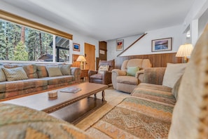 Una sala de estar con sofá, sillones, una mesa de centro y chimenea. Se ven árboles a través de las ventanas.