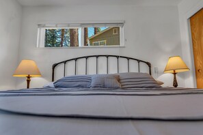A bed with two bedside lamps, a window with a view of trees and a building, and a wooden door. - Walk to Beach | Pet-Friendly | Deck | BBQ (Zephyr Cove)
