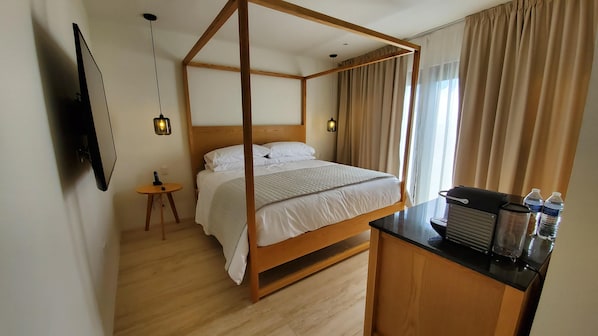 Una cama con dosel de madera y ropa de cama blanca, una mesita redonda con un control remoto, un escritorio con una silla y una botella de agua, y un televisor montado en la pared.