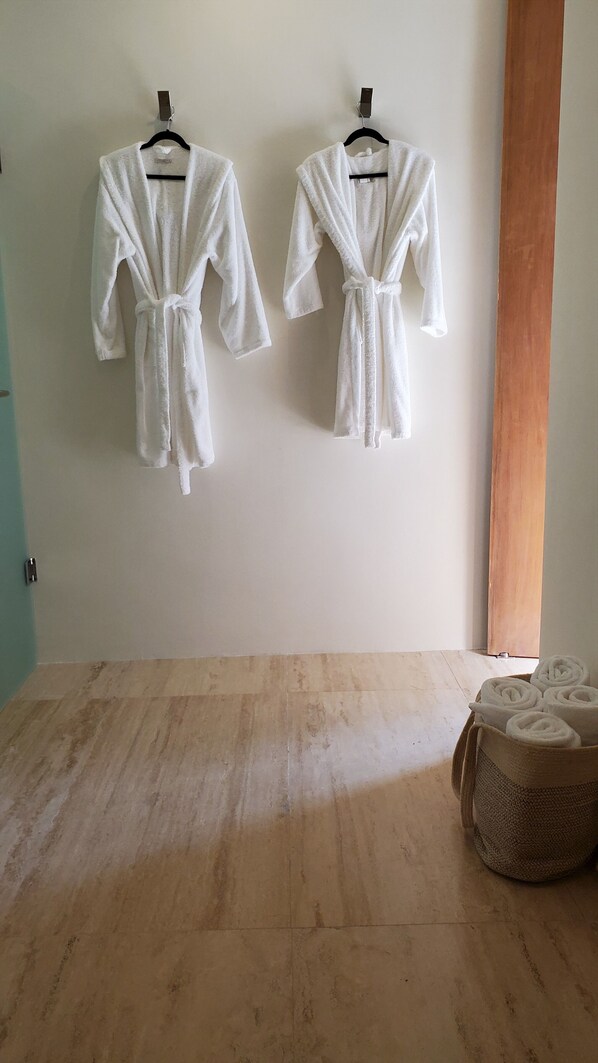 Dos batas blancas colgadas en una pared, suelo de madera y una cesta tejida con toallas enrolladas.