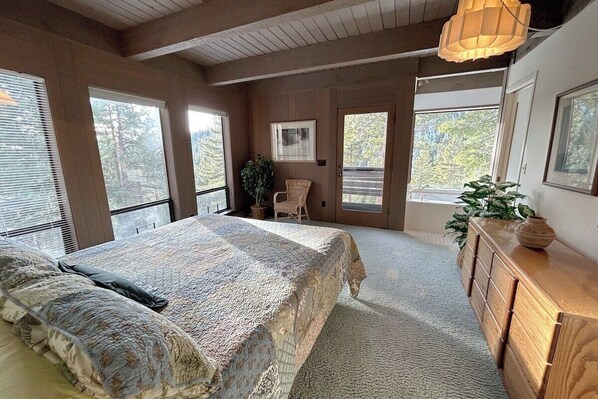 Un dormitorio con una cama grande, un aparador de madera y una silla. Varias ventanas ofrecen luz natural y vistas a árboles.