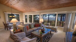 A spacious living room with a wooden ceiling, large windows offering mountain views, a dining area with a table and chairs, and a cozy seating area with a sofa and armchairs. - Hot Tub | Lake Views | Poker | Deck | BBQ (South Lake Tahoe)