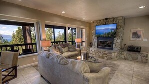 A spacious living room with a large gray sectional sofa, a stone fireplace, and a television mounted on a stone wall. - Hot Tub | Lake Views | Poker | Deck | BBQ (South Lake Tahoe)