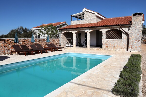 Una piscina con sillones de descanso, un edificio de piedra con techo de tejas rojas y un cielo azul despejado.