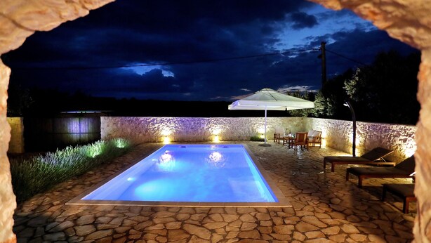 Una piscina de piedra con una sombrilla blanca, rodeada de sillas de descanso y un muro de piedra.
