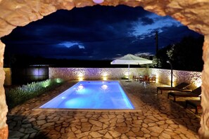 Una piscina de piedra con una sombrilla blanca, rodeada de sillas de descanso y un muro de piedra.