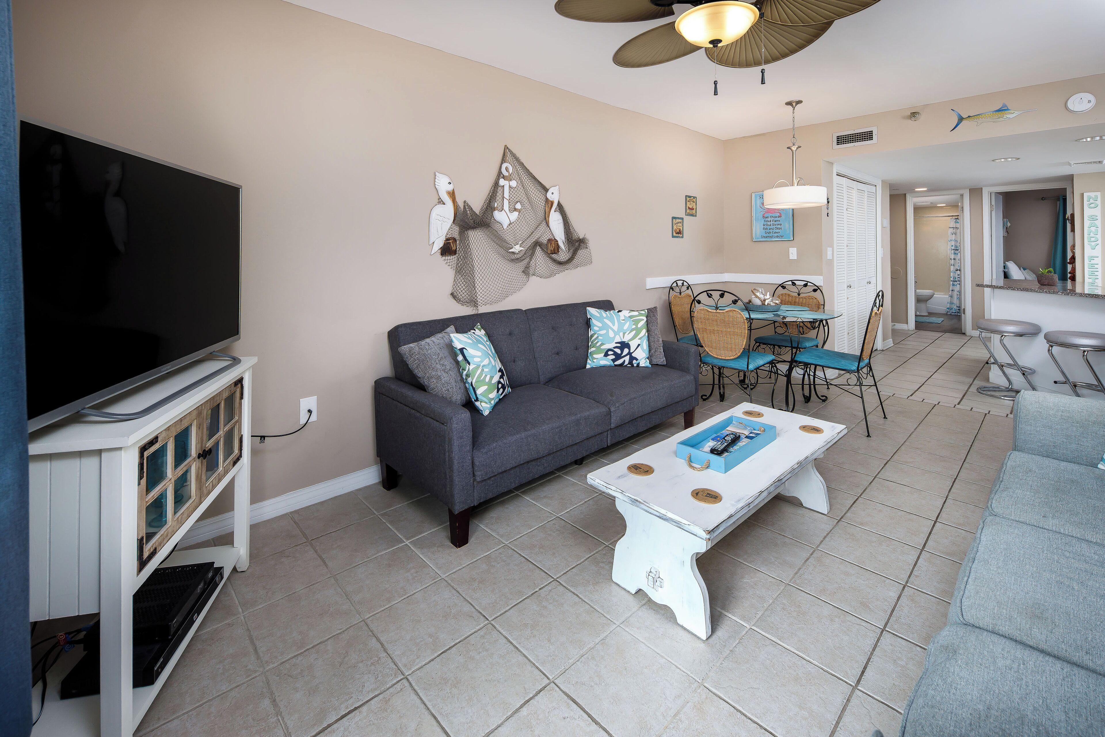 A living room with a grey sofa, a white coffee table, and a TV on a stand. There is a dining area with a table and chairs.