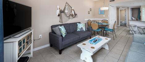 A living room with a grey sofa, a white coffee table, and a TV on a stand. There is a dining area with a table and chairs.