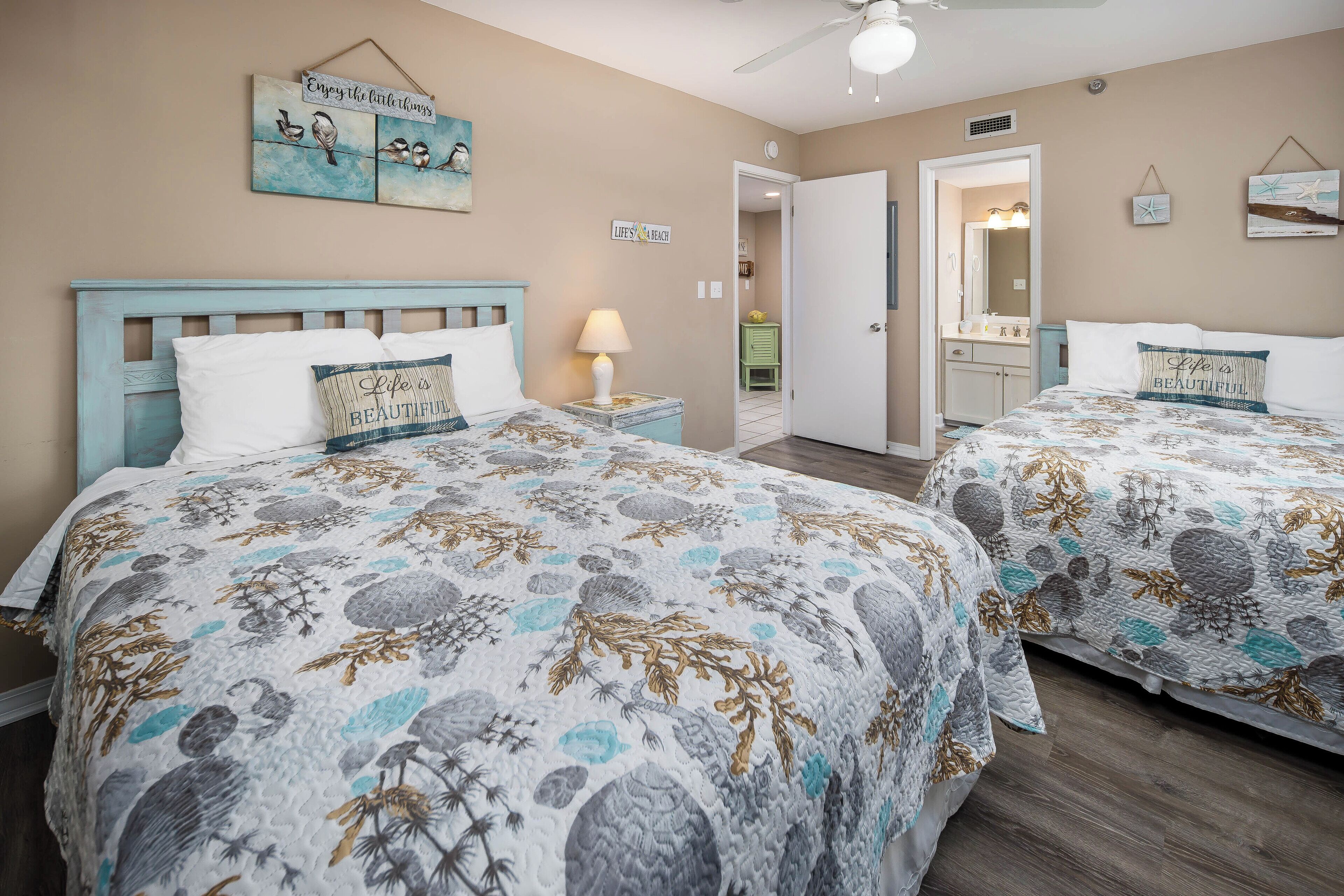 A bedroom with two beds, a nightstand with a lamp, a ceiling fan, and wall decorations including a sign that says "Enjoy the littlerings."