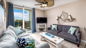 A living room with a grey sofa, a white coffee table, and a grey couch with patterned cushions. A flat-screen TV is mounted on the wall, and there is a ceiling fan above. - Nautilus 2310  Gulf View 2BD/2BA 3rd Floor.  Free Beach Service 3/1-10/31 (Ft Walton Beach)