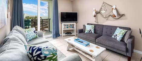 A living room with a grey sofa, a white coffee table, and a grey couch with patterned cushions. A flat-screen TV is mounted on the wall, and there is a ceiling fan above.