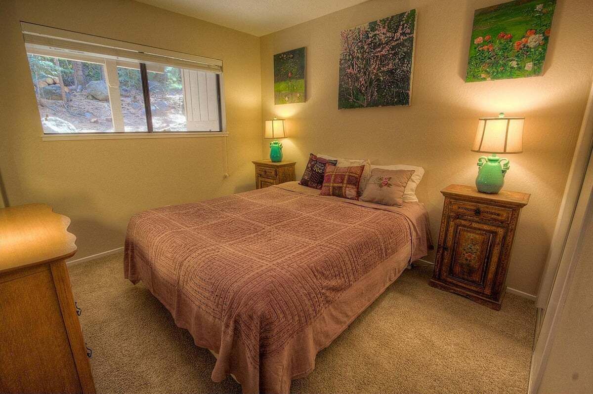 A bedroom with a large bed, two bedside tables with lamps, a window with a view of trees, and two wall-mounted paintings.