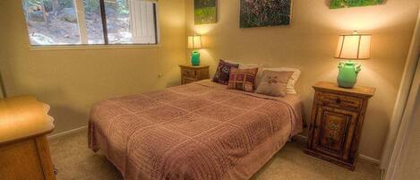 A bedroom with a large bed, two bedside tables with lamps, a window with a view of trees, and two wall-mounted paintings.
