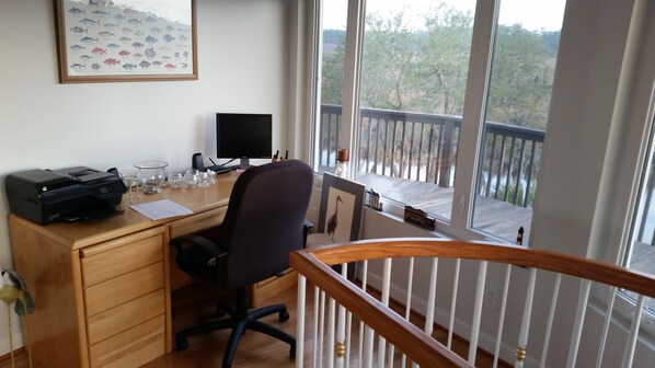 Escritório em casa com mesa, computador e impressora. Há uma janela com vista para árvores e uma varanda.