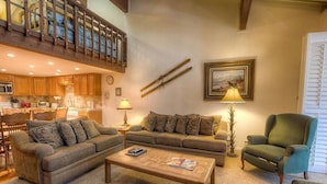 Una sala de estar con sofá, sillón y mesa de centro, un comedor con armarios de madera, y una escalera que lleva a un nivel superior.