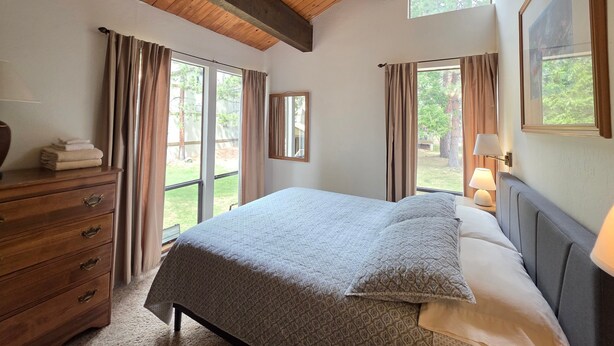A bedroom with a bed, wooden dresser, large windows, and a view of the outdoors. - Kingswood Condo | Community Pool (Kings Beach)